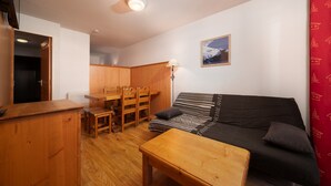 Een knus woongedeelte met een houten eettafel, stoelen, een zwarte bank met gekleurde kussens en een rood gordijn.