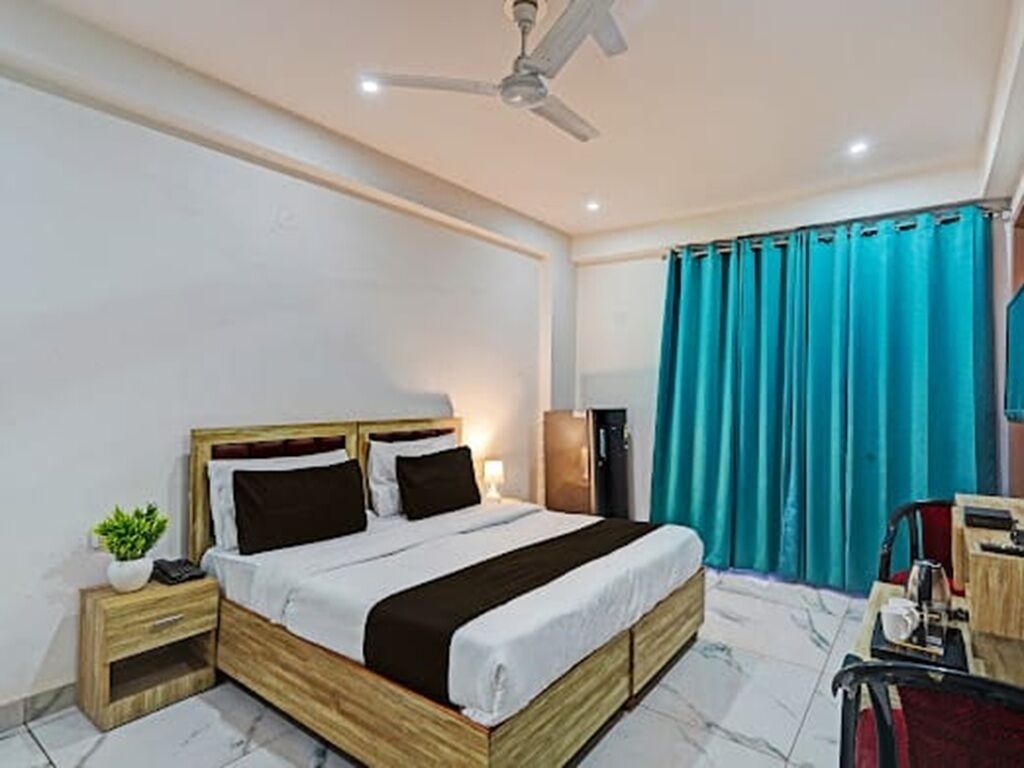 Habitación de hotel con cama, ventilador de techo, mesita de noche con una planta, escritorio con silla y ventana con cortinas.