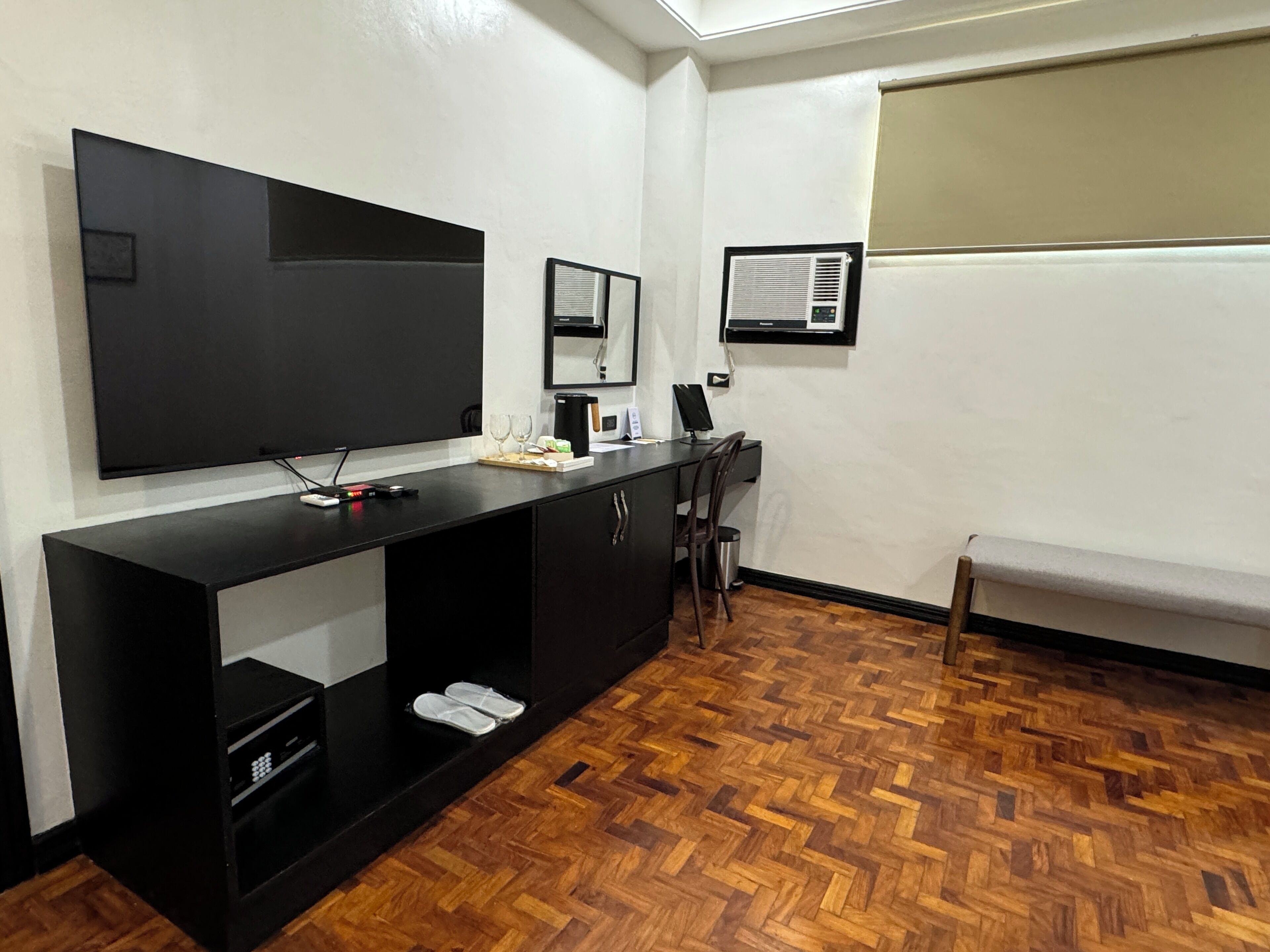 Quarto moderno com uma grande televisão de tela plana na parede, uma escrivaninha preta com um computador e um pequeno espaço de estar com um banco.