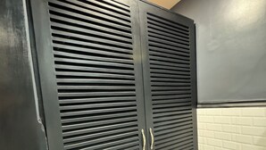 A room with a dark-colored wardrobe with horizontal slats and two handles, a white tiled floor with green accents, and a white brick wall.