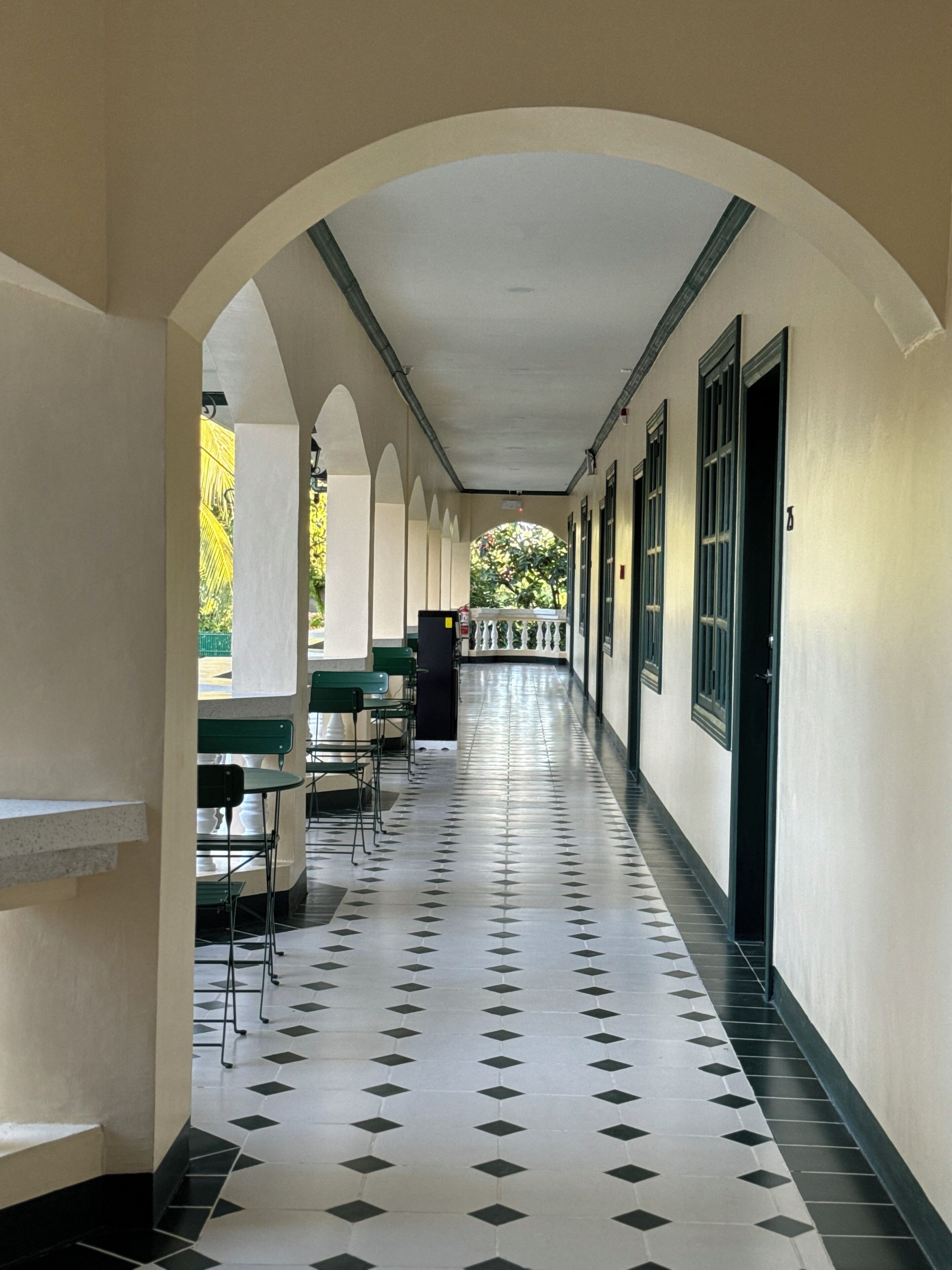 A covered corridor with a tiled floor, green chairs, and tables, leading to a door.