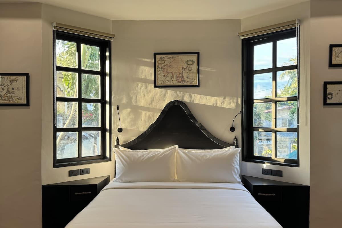A neatly made bed in a room with large windows, a framed map on the wall, and a wooden floor.