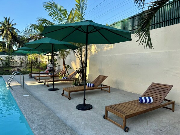 Zona junto a la piscina con sillas de descanso y sombrillas, un patio de concreto y una palmera.