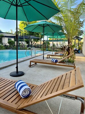 Zona junto a la piscina con sillones de madera, sombrillas verdes y una vista despejada del agua.