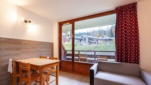 A room with a wooden dining table, chairs, a sofa, and a large window with a view of buildings and a hill. - Apartment in Morillon with Pool & Mountain Views (Morillon)