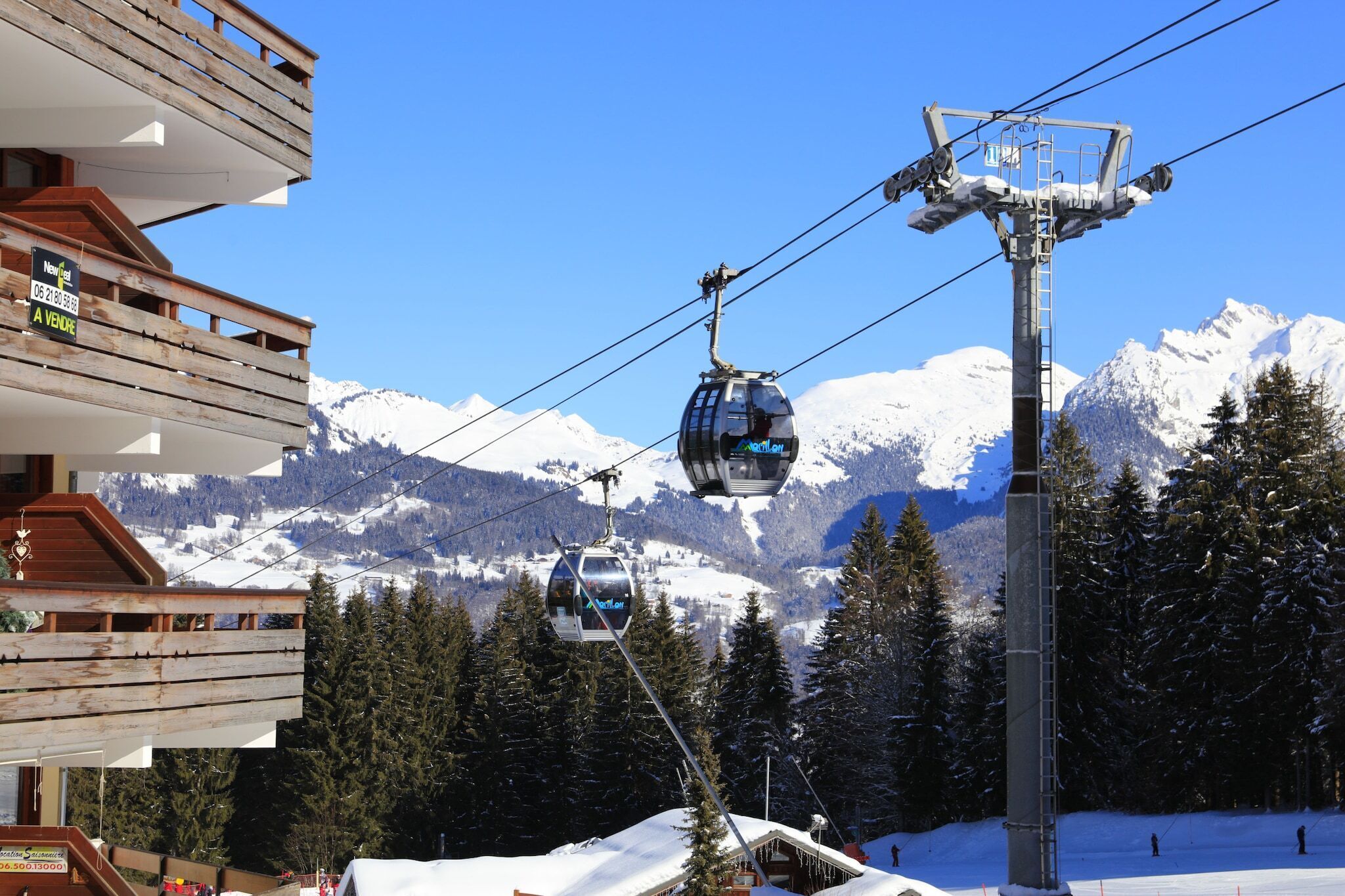 Eine Seilbahn mit Gondeln, ein Holzbalkon und verschneite Berggipfel im Hintergrund.