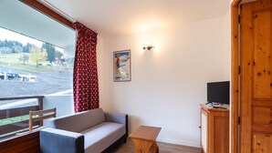 A room with a sofa, a small wooden table, a TV on a wooden cabinet, a poster on the wall, and a window with a view of a landscape. - Apartment in Morillon with Pool & Mountain Views (Morillon)