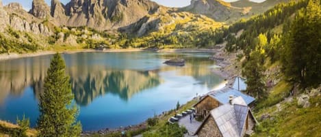 Uma cabana rústica com telhado inclinado, localizada à beira de um lago tranquilo cercado por montanhas.