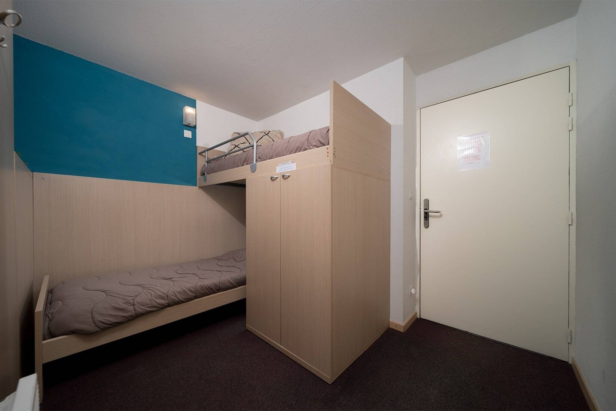 Une chambre de type dortoir, avec un lit superposé, un lit simple, une armoire et une porte.