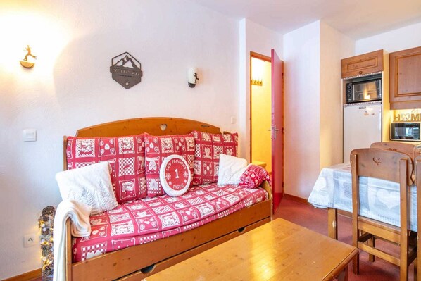 A cozy room with a wooden sofa bed, a small table, and a kitchenette area with a refrigerator and oven.