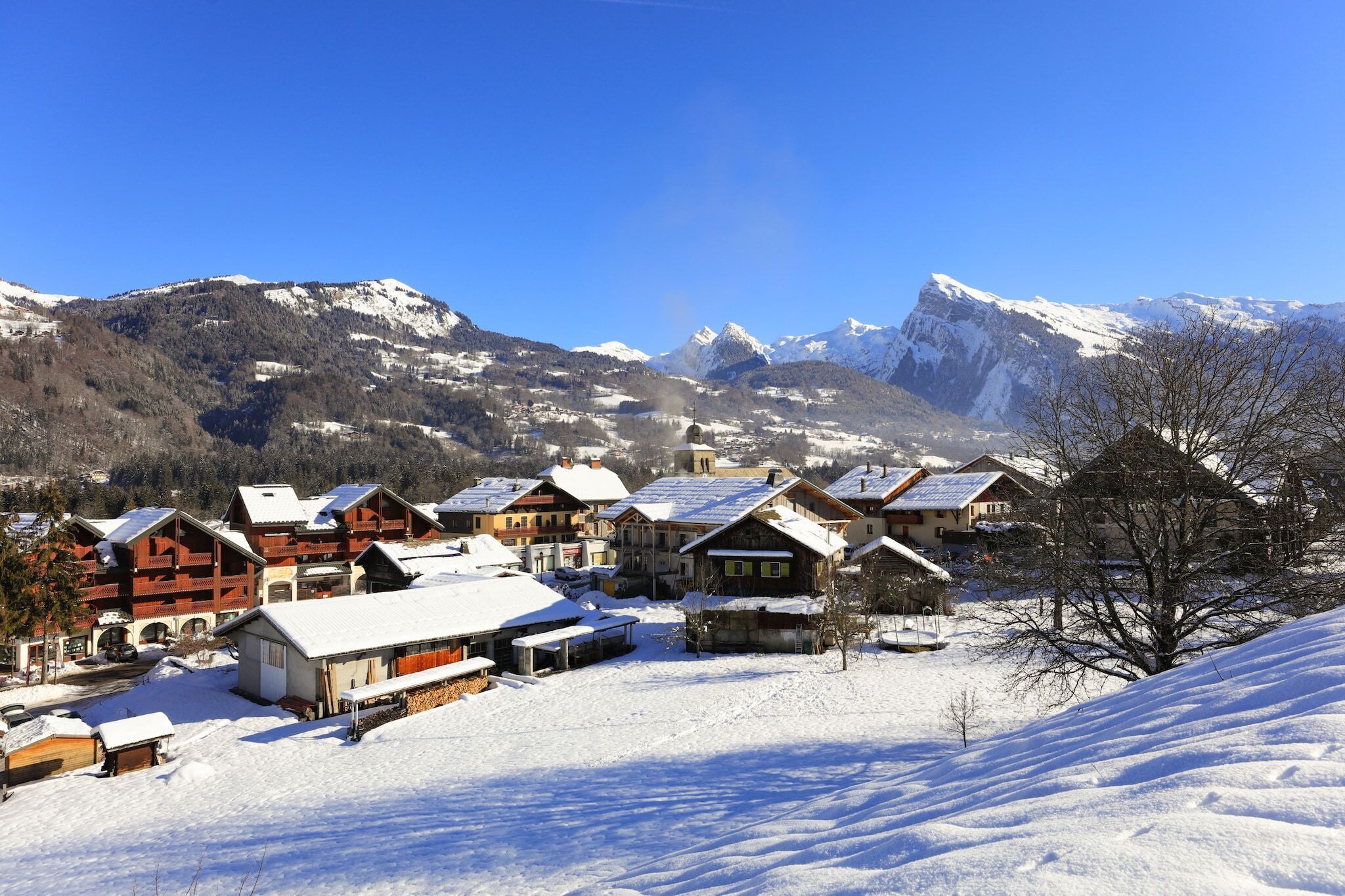 A snow-covered village with traditional wooden houses, a clear blue sky, and snow-capped mountains in the background.