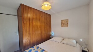Ein Schlafzimmer mit einem Holzschrank, einem Bett mit weißer Bettwäsche, einem Nachttisch mit Lampe und einem an der Wand befestigten Spiegel.