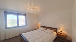 A bedroom with a large bed, two bedside tables with lamps, a window with a view of buildings, and a ceiling light fixture.