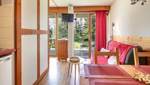 A cozy room with a wooden floor, a dining table with placemats, a sofa with a red blanket, and a view of a deck and trees outside. - Terrace Apartment in Chamrousse (Chamrousse)