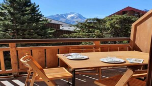 A wooden deck with a table and chairs, overlooking a mountain and a building with a red roof.