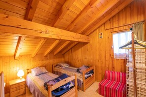 Una habitación tipo cabaña de madera con dos camas individuales, una mesita con una lámpara, un sofá a rayas rojas y una ventana con cortinas.