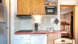 A compact kitchen with wooden cabinets, a built-in oven, a dishwasher, and a small dining area with a chair. - Alpine Apartment with Terrace (Chamrousse)