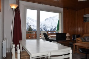 A room with a table, chairs, a TV, and a view of snow-covered mountains through large windows.