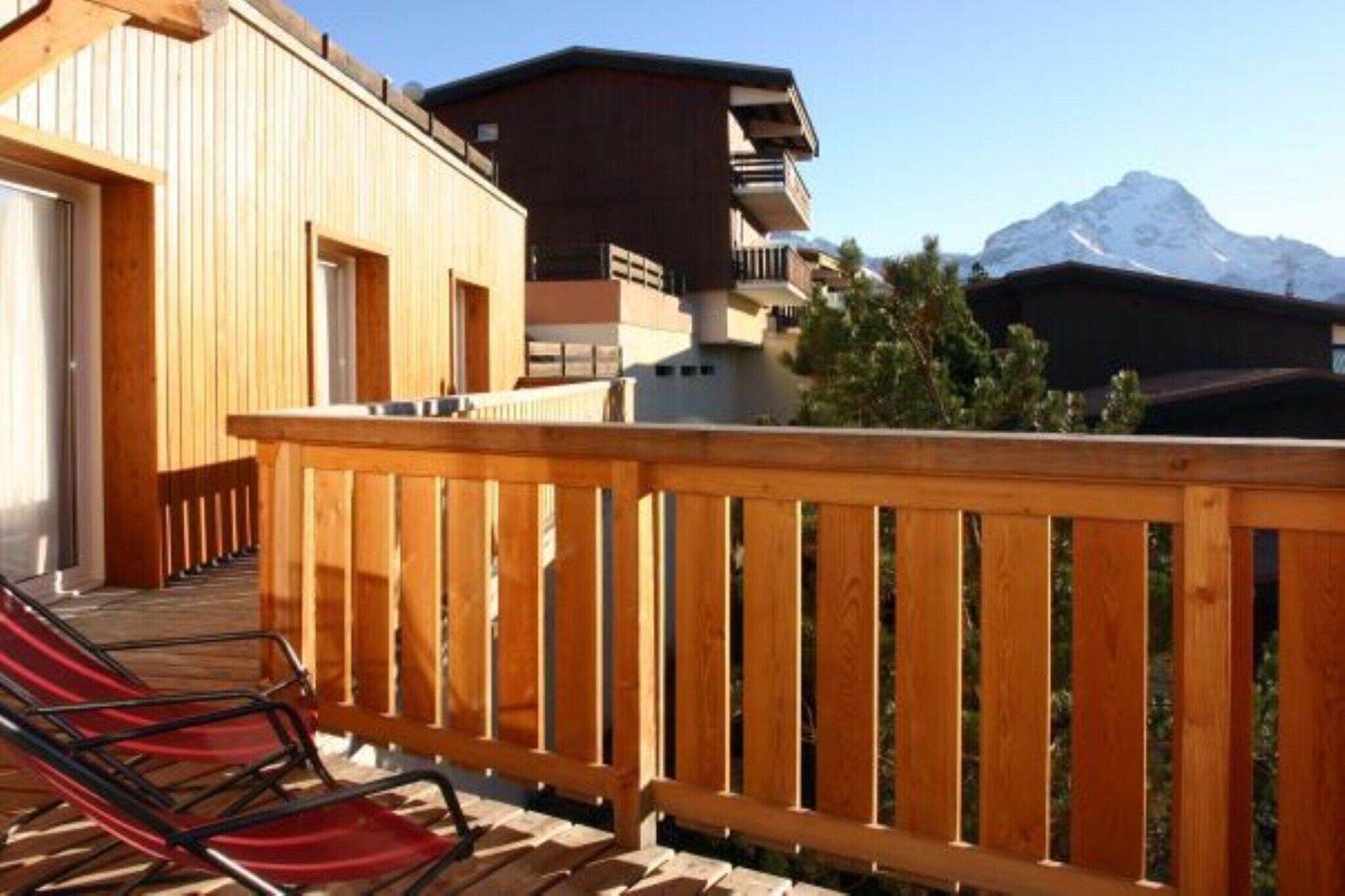 A wooden deck with a red chair, a mountain view, and a building with balconies.