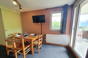 Uma sala de jantar com uma mesa de madeira, cadeiras, uma TV fixada na parede, uma janela com cortinas e um radiador.