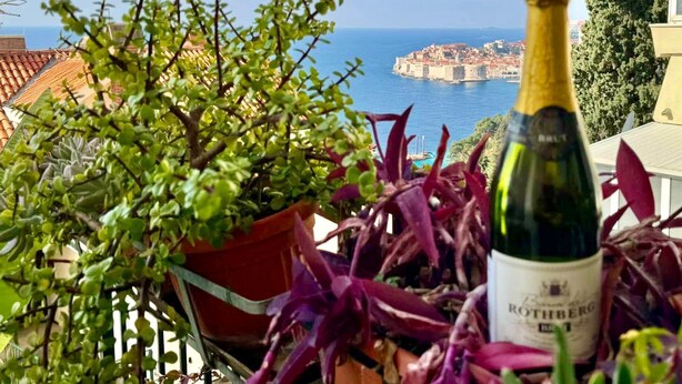 Uma garrafa de vinho numa varanda com vista para uma cidade costeira e uma fortaleza histórica.