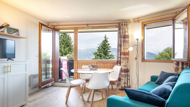 A cozy room with a teal sofa, a dining table with chairs, a TV, and a view of trees and mountains through large windows.