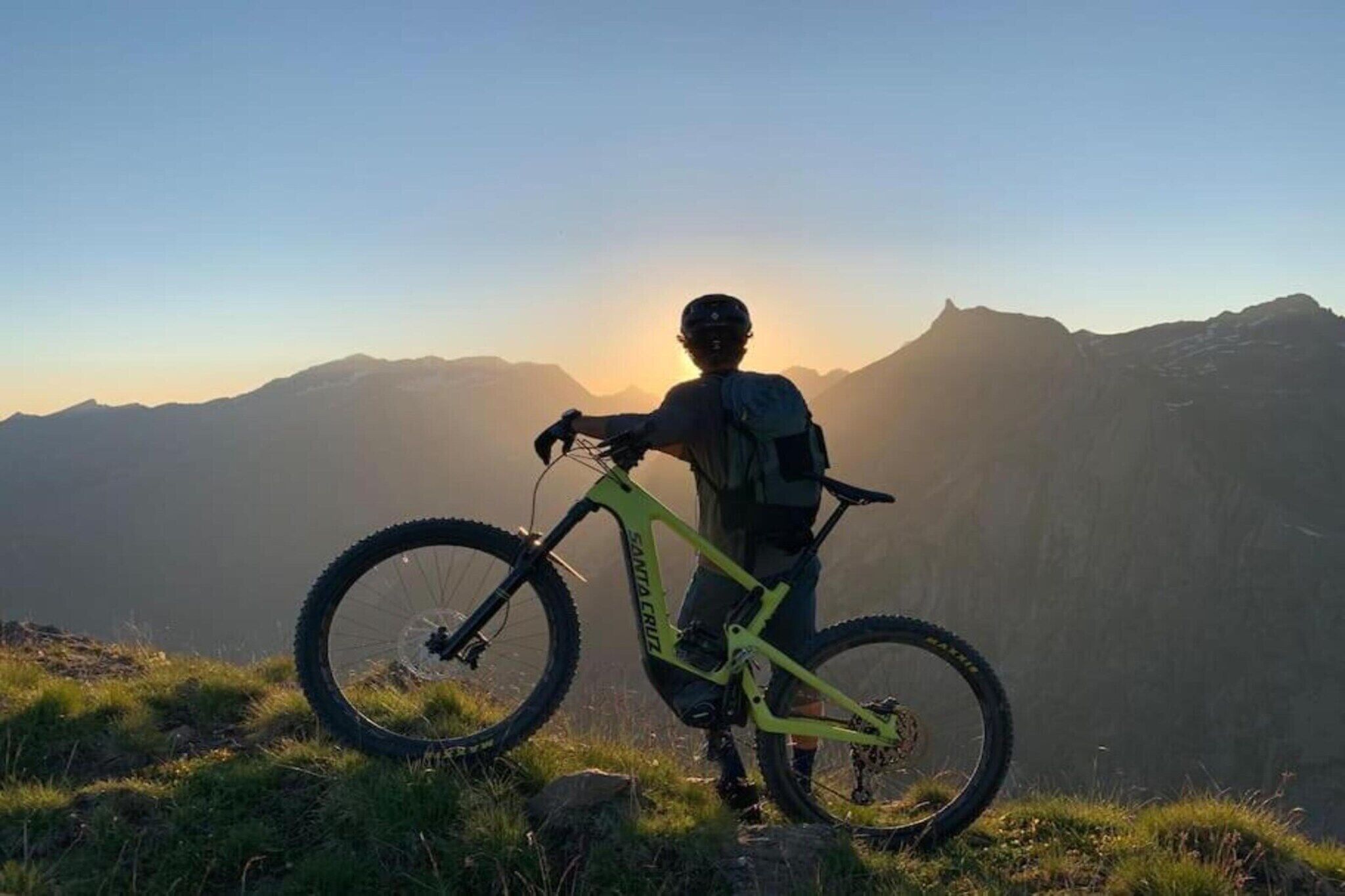 A person standing on a grassy hill with a mountain bike, overlooking a mountainous landscape.
