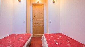 A narrow room with two single beds, each with a red bedspread, a wooden door, and two wall-mounted light fixtures. - Apartment in Orcières with Heated Pool (Orcières)