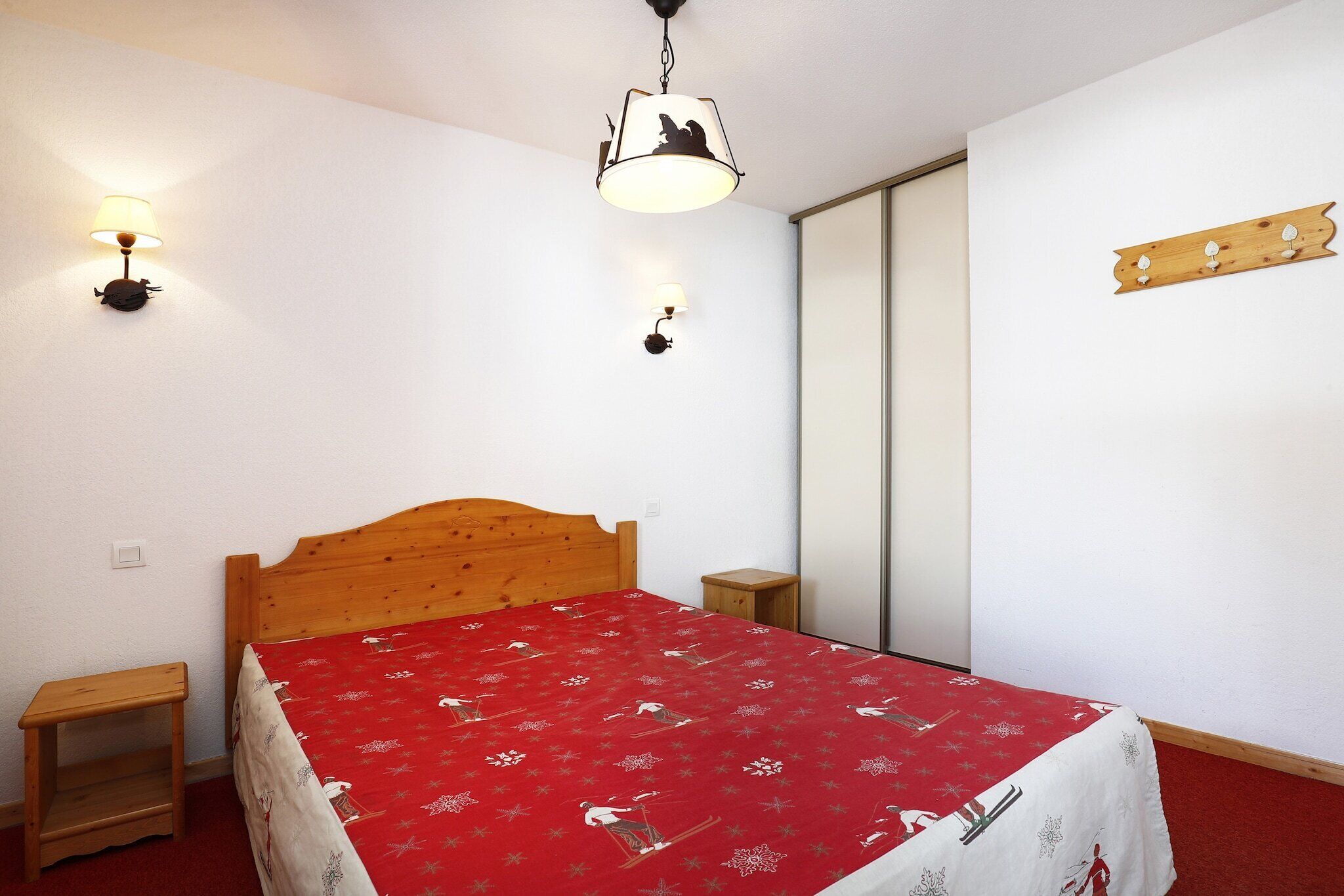 Quarto com cama de madeira, colcha vermelha, duas mesas de cabeceira, luminária fixada na parede e prateleira de madeira.