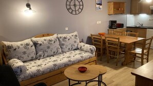 Un salon confortable avec un canapé, une table à manger et des chaises. Une horloge est fixée au mur, et on aperçoit un coin cuisine à l’arrière-plan.