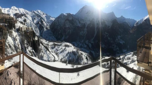 Un paysage montagneux enneigé, avec un ciel dégagé et un soleil brillant derrière les montagnes.