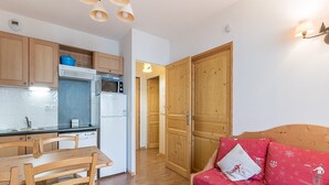 Une cuisine compacte avec des meubles en bois, un four à micro-ondes et un réfrigérateur. On y trouve une table à manger avec des chaises, ainsi qu’un canapé rouge agrémenté d’un coussin sur le thème du ski.
