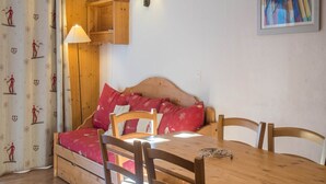 Ein gemütliches Zimmer mit einem hölzernen Bett, einem Esstisch und Stühlen. Auf dem Tisch steht eine Lampe und über dem Bett befindet sich ein Wandregal.