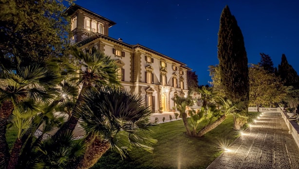 A large, well-lit building with a symmetrical facade, surrounded by lush greenery and a stone pathway leading to the entrance.