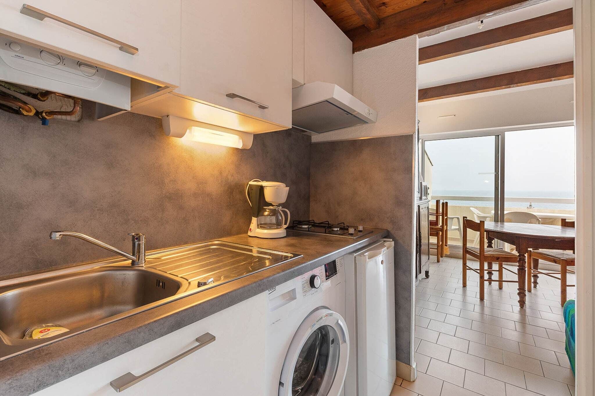Une cuisine compacte équipée d’un lave-linge intégré, d’une machine à café et offrant une vue sur la plage par la porte ouverte.
