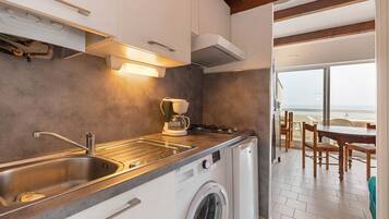 Une cuisine compacte équipée d’un lave-linge intégré, d’une machine à café et offrant une vue sur la plage par la porte ouverte.