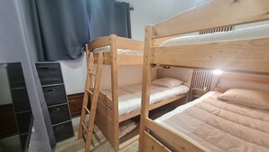 Een slaapkamer met een stapelbed, een houten vloer, een klein bureau met lades en een raam met gordijnen.
