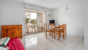 A room with a bed, a wooden cabinet, a dining table with chairs, and a sliding glass door leading to an outdoor area with chairs. - Charming Coastal Escape in Agde (Agde)