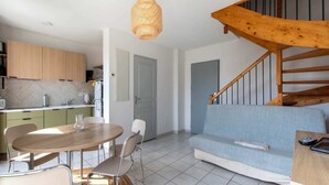 A modern kitchen with wooden cabinets, a dining table with chairs, a blue sofa, and a wooden staircase. - Modern Seaside Apartment (Le Grau-du-Roi)