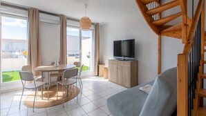 A modern living room with a sofa, a dining table with chairs, a TV on a wooden cabinet, and a staircase. - Modern Seaside Apartment (Le Grau-du-Roi)