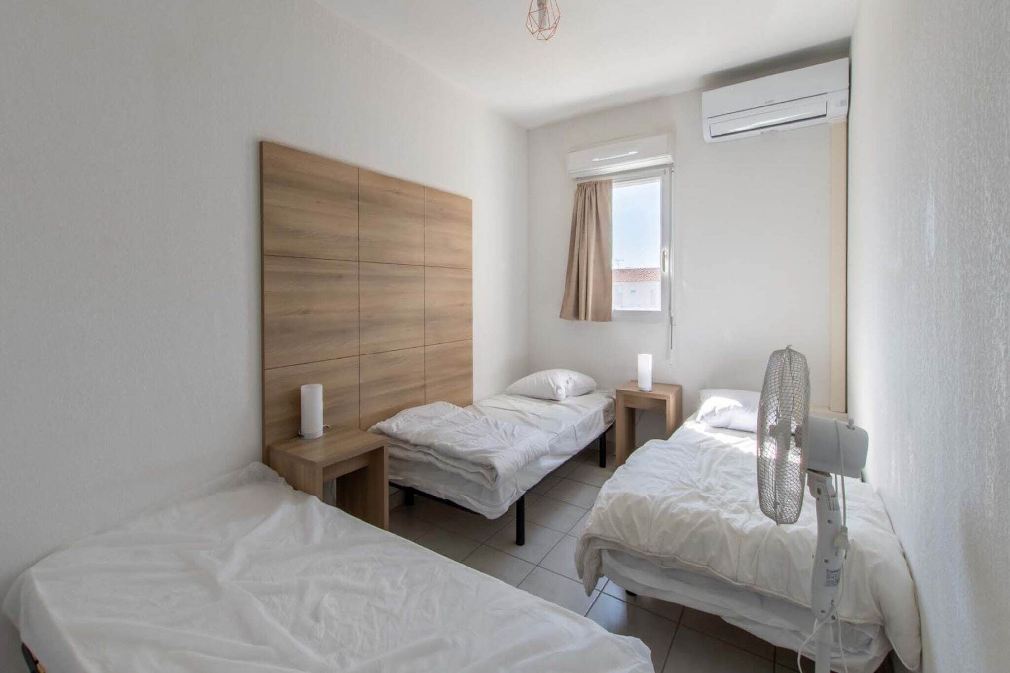Une chambre d’hôtel avec deux lits simples, une tête de lit en bois, une fenêtre avec des rideaux, un appareil de climatisation et un ventilateur sur pied.