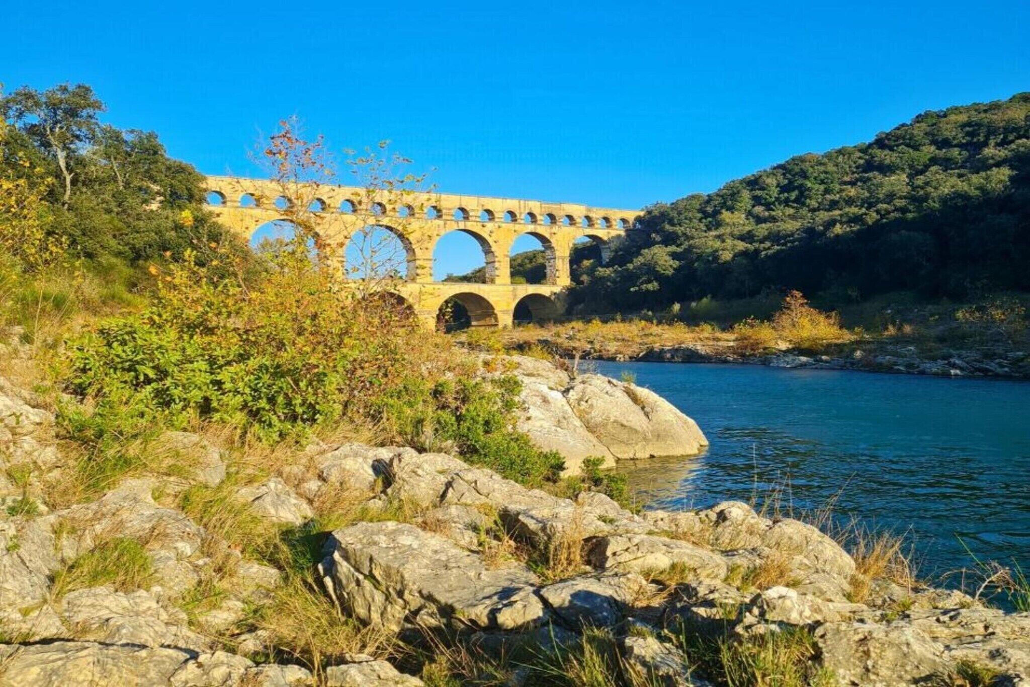Eine steinerne Brücke, ein Aquädukt, führt über einen Fluss mit felsigem Gelände und viel Grün.