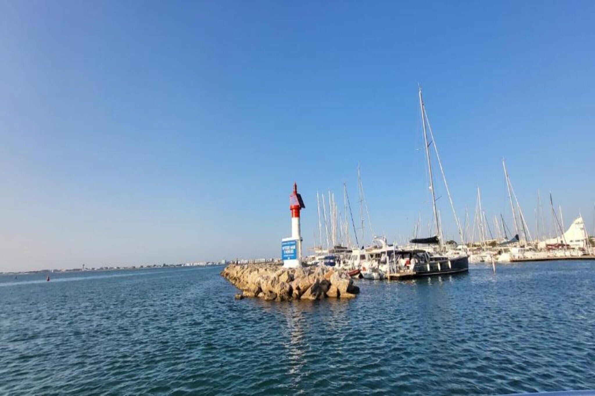 Een jachthaven met een vuurtoren op een rotsachtige landtong, omgeven door kalm water en tal van zeilboten.