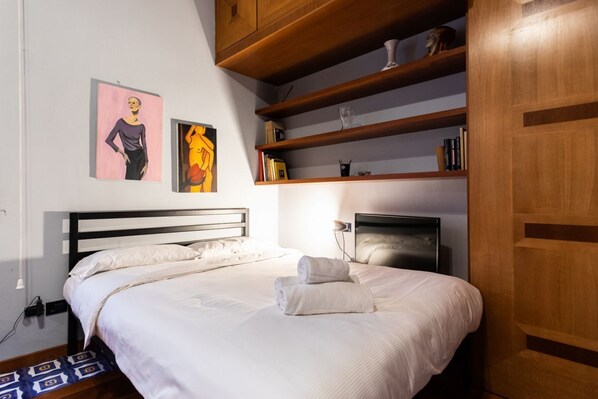 Una cama bien tendida con sábanas blancas y dos toallas enrolladas, un cabecero de madera, y una repisa con libros y objetos decorativos.