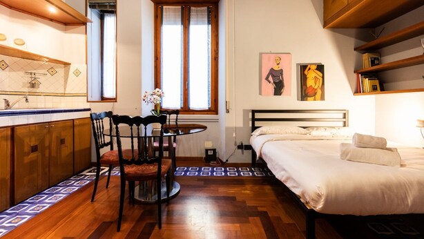 Une chambre d’hôtel de taille réduite, comprenant un lit, une petite table avec des chaises, une fenêtre avec des rideaux, une armoire en bois et un mur recouvert de carreaux.
