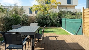Une terrasse avec une table et des chaises, une plateforme en bois et une vue sur un bâtiment moderne, avec un arbre au premier plan.