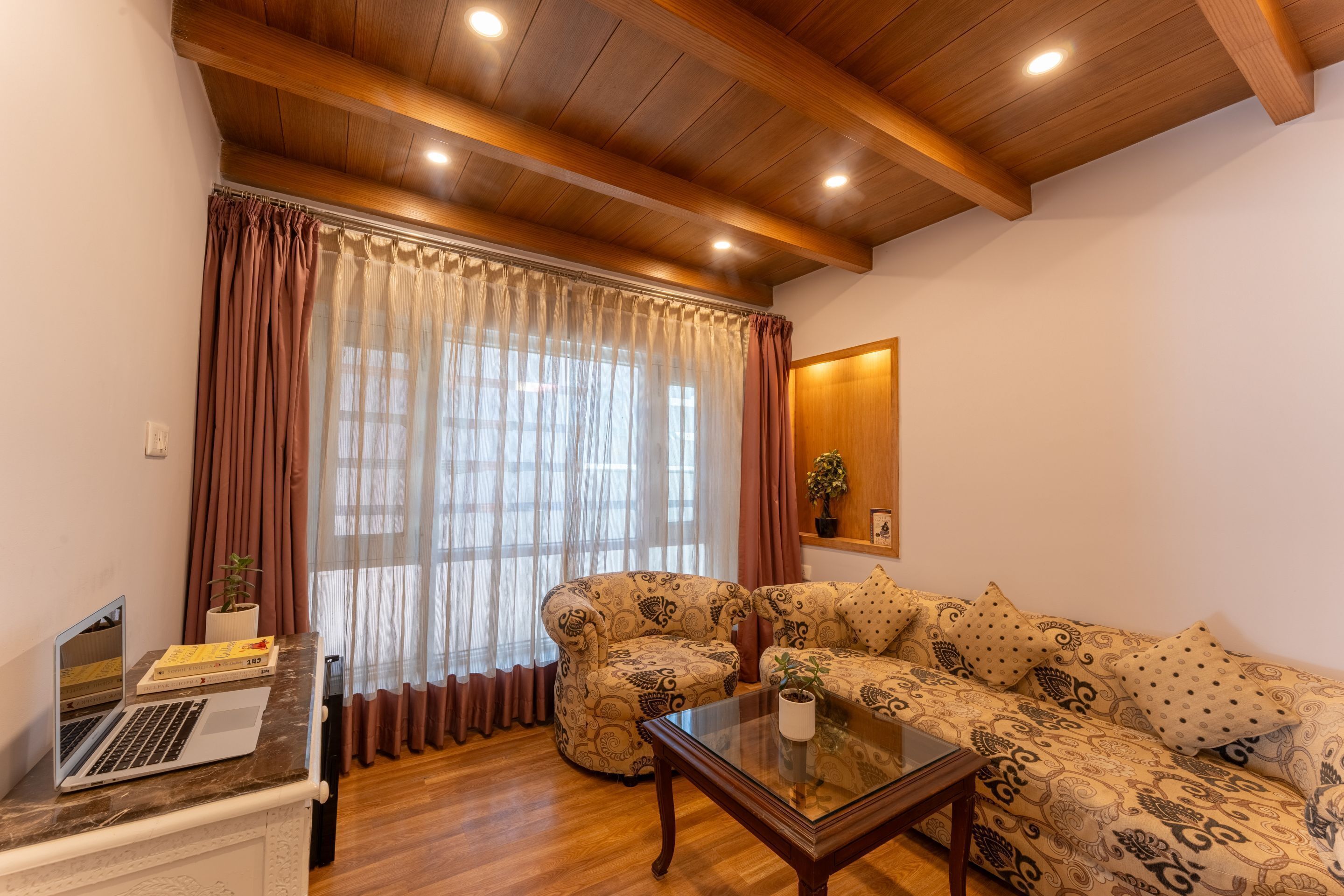 Sala de estar com teto de madeira, sofá, mesa de centro de vidro e um laptop sobre uma mesinha lateral.