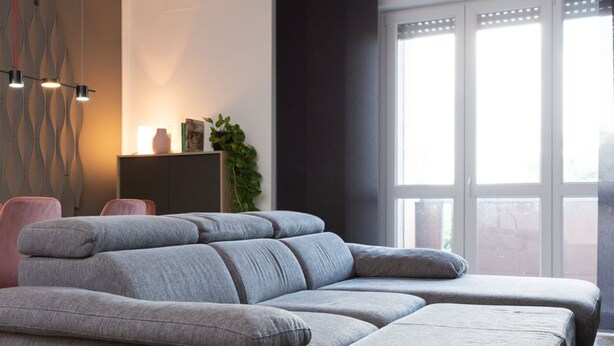 A modern living room with a grey sectional sofa, a side table with a vase and books, and a large window with blinds. - Exclusive Guest House - Fiera Milano Rho (Rho)