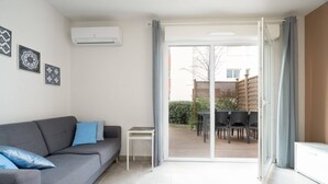 A modern living room with a grey sofa, a small white table, and a sliding glass door leading to an outdoor patio with a table and chairs. - Cozy Villa Steps from the Beach (Valras-Plage)