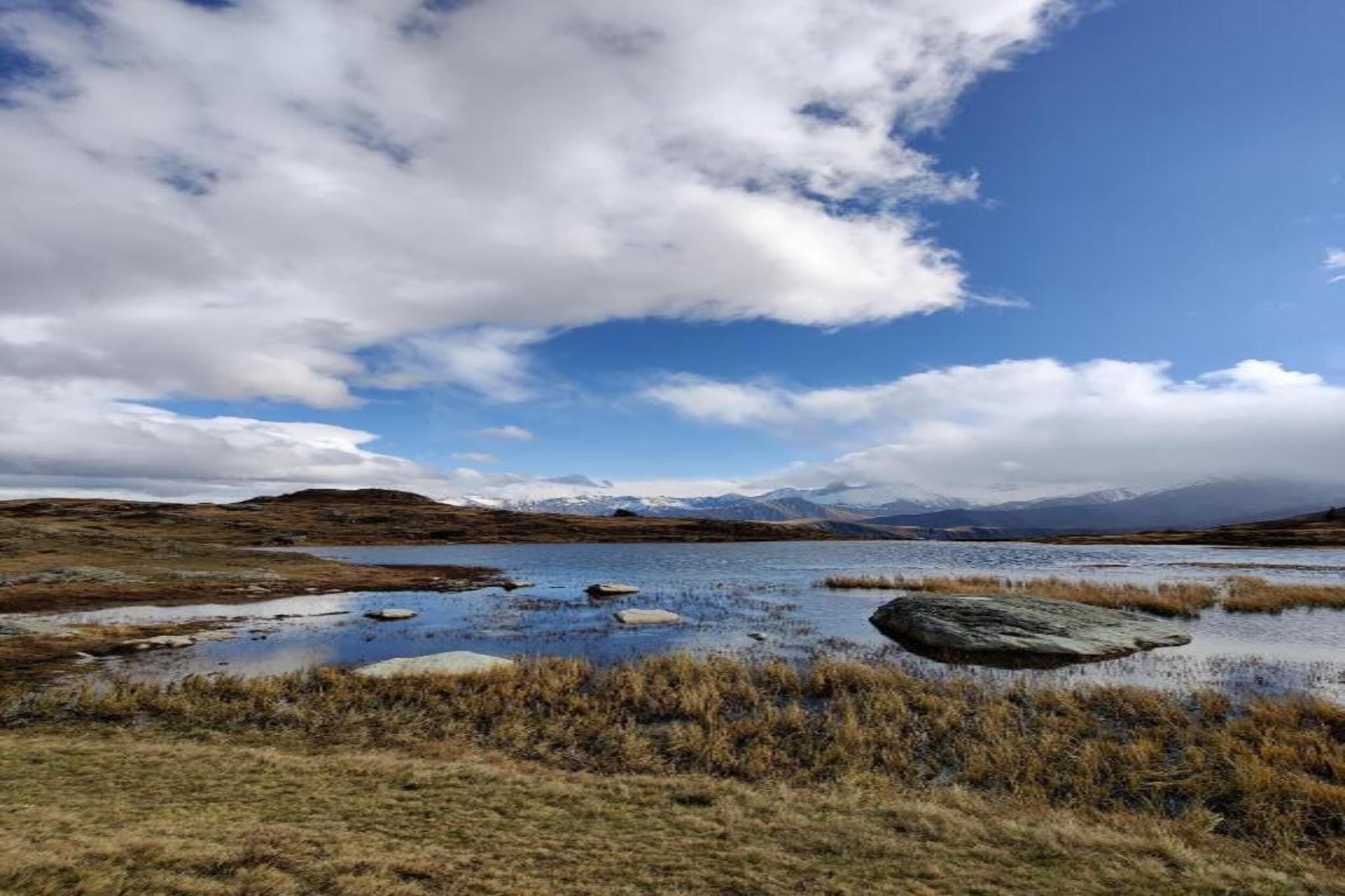 A natural landscape with a lake, grassy areas, and distant mountains under a partly cloudy sky.